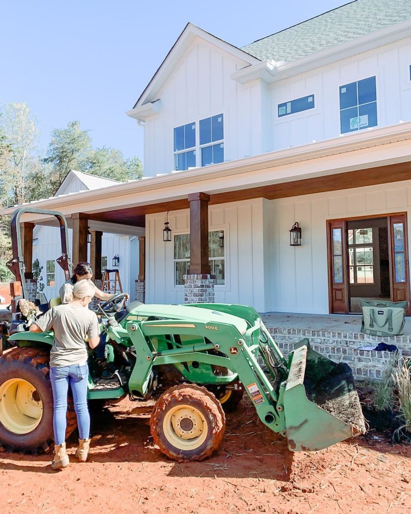 Fawn of Those Plant Ladies teaching a crew member to use the John Deere tractor in front of a white farmhouse.